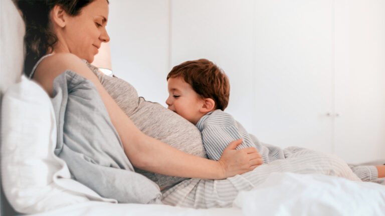 Mujer embarazada sentada en la cama con su hijo apoyado en su vientre.