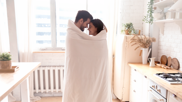 Pareja abrazándose de pie, tapados con una colcha.